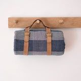 Plaid fabric roll-up bag with brown straps hanging on a wooden rack against a white background