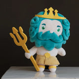 A plush toy character resembling a sea king, with turquoise blue curly hair and beard, a white face and body, a yellow crown, and a yellow trident, standing on a white surface against a dark background.