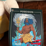A Poseidon card or illustration featuring an illustrated sea god with blue hair, a trident, and a yellow skirt, photographed in a casual indoor setting with a plush toy and brown fabric in the background.