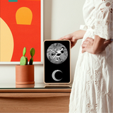 A person in a white lace dress touching a wood clock with a circular screen showing a black-and-white cartoon character. The scene is styled in a modern room with a glass-topped table, a small potted cactus, and a colorful abstract painting.