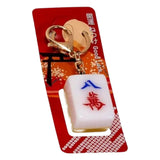 A white mahjong tile keychain with red and blue characters, attached to a gold clasp and displayed on a red Good Luck card.