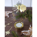 A gold metal tealight candle holder with star and windmill details, displayed on a dark wooden surface with a lit tealight candle, with pine branches and gingerbread cookies in the background.