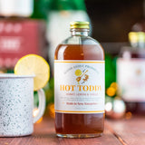 Bottle of 'Hot Toddy' with a label on a wooden surface, surrounded by lemons and mugs.