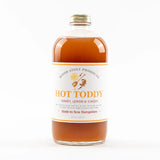 A clear glass bottle filled with amber-brown liquid, sealed with a gold cap, and labeled 'HOT TODDY' by Wood Stove Products. The white label includes orange and brown text and graphics, and the bottle is photographed against a clean white background.