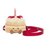A plush cake-shaped crossbody bag with cream-colored frosting, red berries, a red and white striped candle, and a red strap, shown on a plain white background.