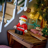 A LEGO Santa Claus figure stands on a wooden table in a decorated holiday setting with wrapped gifts, a Christmas tree, and staircase railings in the background.