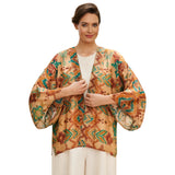 A woman modeling a loose, open-front kimono jacket with 3/4 sleeves, featuring a warm tan base with brown, teal, and green geometric and floral patterns, worn over a white top and white pants against a plain white background.