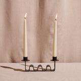 A wavy black metal candle holder with two lit taper candles placed in it.