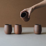 A set of four handcrafted brown ceramic cups with a matte finish on a plain background. A hand is holding one of the cups
