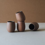 A set of four handcrafted brown ceramic cups with a matte finish on a plain background.