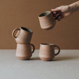 A set of four beige ceramic mugs with a handle, placed on a neutral surface.