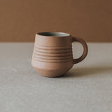 A beige ceramic mug with a handle, placed on a neutral surface.