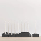 A modern black geometric candle holder set with nine white taper candles, displayed on a light wood surface against a clean white background.