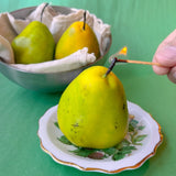 Yellow pear candle on a decorative plate with a hand holding a lit match, against a green background.
