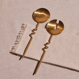 A set of two brass finish servers with a spiral design, on a neutral fabric background.