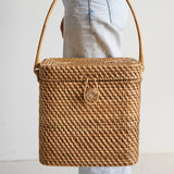 Woven picnic basket with a handle held by a person wearing light-colored pants on a neutral background