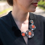 A close-up lifestyle shot of a woman wearing a black V-neck top and a chunky statement necklace. The necklace features black geometric fabric discs decorated with orange-red floral patterns on a white background.