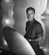 A black and white portrait of a man standing indoors, wearing a short-sleeve checkered shirt and dark trousers, with large sculptural ribbed lamps visible behind him.