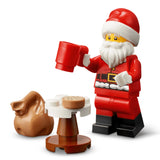 A LEGO Santa Claus minifigure holding a red mug, standing next to a small white and brown table with a cookie and a brown sack, on a white background.