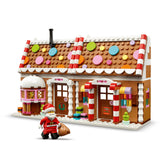 A gingerbread house building set with a Santa minifigure, shown on a clean white background. The house features a brown brick-style facade, white trim, colorful round decorations on the roof, red-and-white striped columns, and a small black chimney.