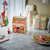 A small gingerbread house building set with a tiny Santa minifigure stands on a marble tabletop. The scene is decorated for Christmas, with wrapped gifts, a plate of cookies, and warm holiday lights in the background.