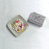 A small square dish filled with multicolored candy pieces next to a grey box labeled KONPEIDOU, photographed on a light neutral background.