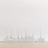 A minimalist white menorah-style candle holder with seven taper candles, displayed on a light wood surface against a clean white background.