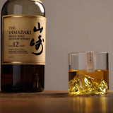 A bottle of Yamazaki 12 Years Single Malt Japanese Whisky stands next to a clear rocks glass filled with amber whisky and a sculpted mountain-shaped interior on a wooden surface.