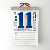 A Japanese calendar hanging on a white wall, featuring a large blue '11' in the center with a pink and white abstract design behind it. The calendar has a silver metal header with a red hanging ribbon and the word 'CALENDAR' on it.