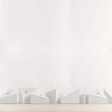 A row of minimalist geometric candle holders in a light gray color, arranged along the bottom edge of a light wood surface against a clean white wall.