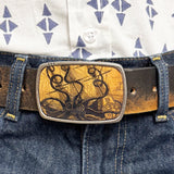 Person wearing a patterned shirt and blue jeans with a decorative belt buckle featuring an octopus attacking a ship.