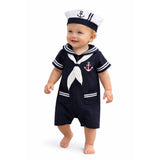 A baby wearing a navy blue and white sailor costume set, including a short-sleeve romper with a white neck scarf and anchor emblem, and a matching white sailor hat standing against a plain white background.