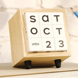 A wooden cube desk calendar with white tiles displaying the days of the week and the date October 23, with 'umbra' printed on one tile. The calendar stands on two small black feet against a light marble-like background.