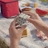 A person's hands holding a wooden letter cube puzzle on a light-colored picnic blanket, with a red cooler bag, watermelon, and juice box in the background.