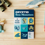 A book titled 'Cryptid Sea Monsters: A Field Guide' lies on a light wood tabletop, surrounded by small props including binoculars, a shark toy, a mermaid doll, and colored pencil.