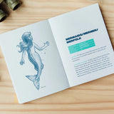 An open children's book on a light wood tabletop, showing a blue mermaid illustration on one page and a mermaid-themed text page on the other. 