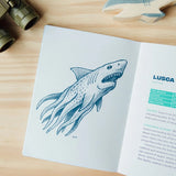 Open book with shark illustration, wooden shark toy, and binoculars on a wooden surface