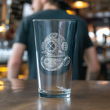 A clear pint glass with a white etched vintage diving helmet design, standing on a wooden bar counter with a blurred bartender in the background.