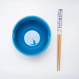 A bright blue bowl shown next to a pair of wooden chopsticks with a blue and white patterned handle, on a plain white background.