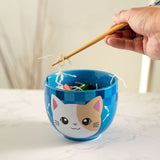 A blue ceramic bowl with a cute cartoon cat face printed on the front, shown on a light marble surface. A hand is holding wooden chopsticks above the bowl, with noodles visible inside.