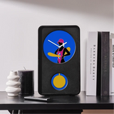 A black rectangular clock with a circular blue face showing a stylized figure in pink and yellow, plus a smaller yellow circular detail below, displayed on a dark tabletop beside books and candles.
