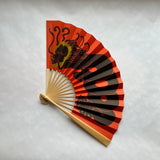 A traditional folding hand fan with a red and black design, featuring a black dragon illustration and gold accents, set against a light gray textured background.