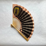 A folding hand fan featuring a black and brown monkey illustration with a yellow-green face on the upper panel, set against a light gray textured background.