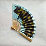 A traditional folding hand fan with a light blue and black ox pattern, featuring a natural bamboo handle on a light gray textured background.
