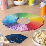 A circular color swatch fan made of overlapping rectangular playing cards arranged in a rainbow spectrum on a wooden table, with a few cards placed in front and snacks nearby in a real setting.