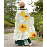 A person standing outdoors on a path, shown from the back, wearing a long white haori with large yellow floral prints and green stems, over a dark green skirt and light patterned hem, with a tan hat.