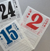 Three small white paper calendar pages with large printed numbers 24, 15, and 2 in black, blue, and red, plus Japanese text and small boxes for writing, arranged on a gray surface.