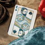 Playing card with a king of spades design on a sandy surface with water and rope.