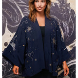 A woman wearing a navy blue open-front kimono-style jacket with embroidered celestial patterns including stars, constellations, and a rabbit motif, posed against a dark abstract painted backdrop.