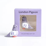 A cute, stylized toy pigeon figurine in shades of lavender purple, white, and bright orange feet, set against a clean light gray background.